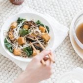 hand with chopsticks over white bowl of vegetables and noodles