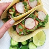 hand holding tacos on white plate with lime and cucumber