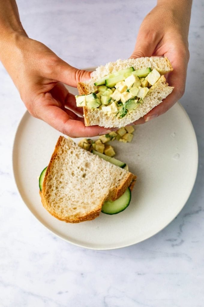 hands holding vegan egg salad sandwich on white plate