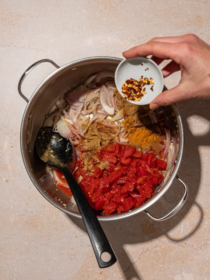 hand adding chile flakes to pot with tomato and onion