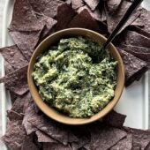 spinach dip in brown bowl with spoon and blue chips on white tray