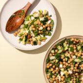Chickpea cucumber salad on white plate with wooden spoon and bowl on yellow background