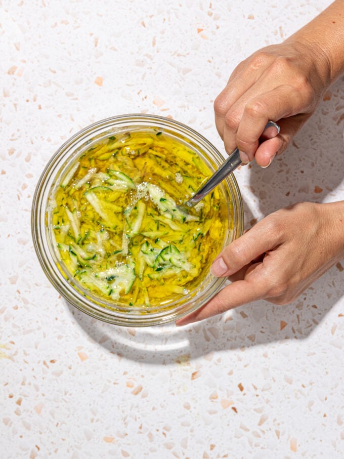 hand stirring grated zucchini and oil in bowl