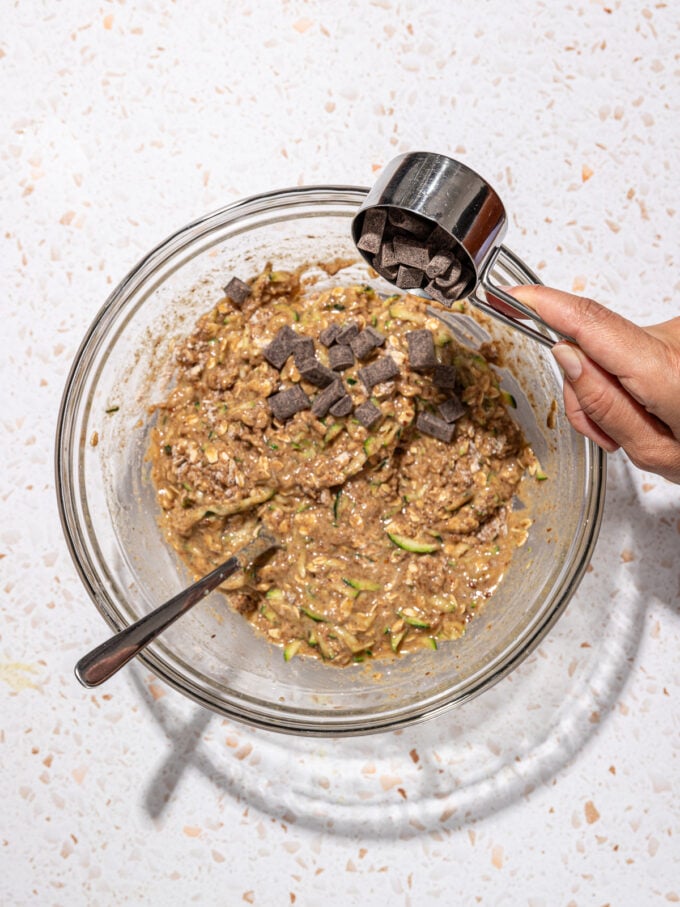 hand adding chocolate chunks to bowl of zucchini muffin batter