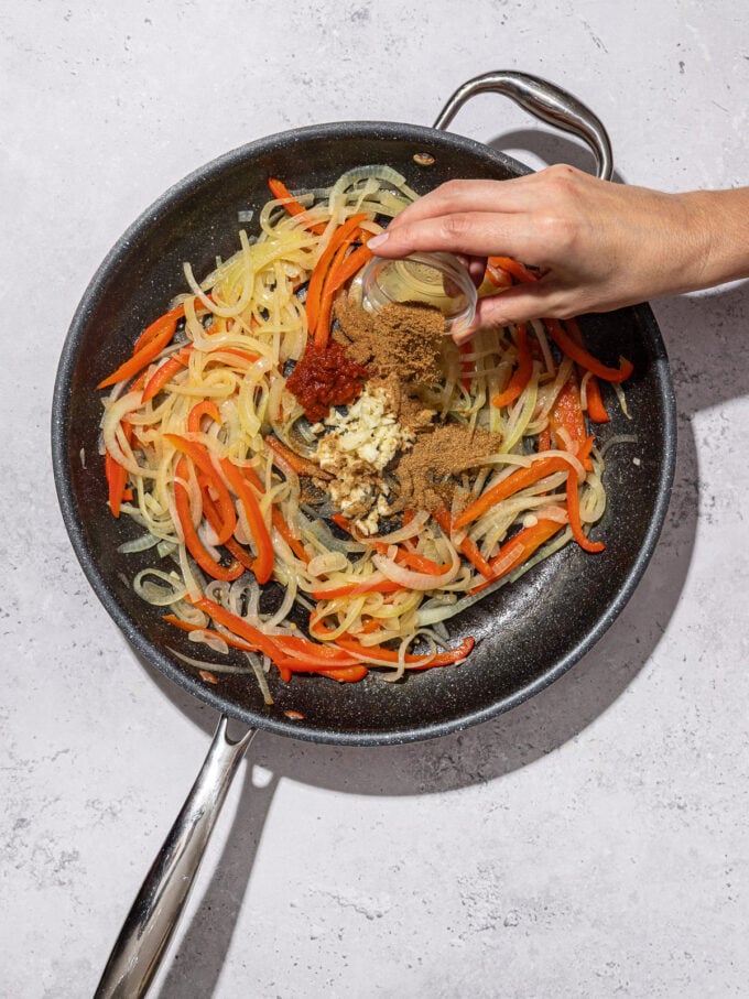 hand adding spices to onion and pepper in skillet
