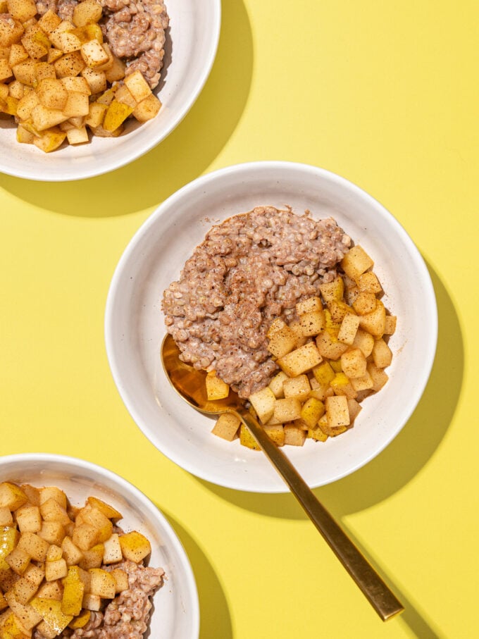 white bowls with buckwheat porridge, diced cooked pears and a gold spoon on yellow background
