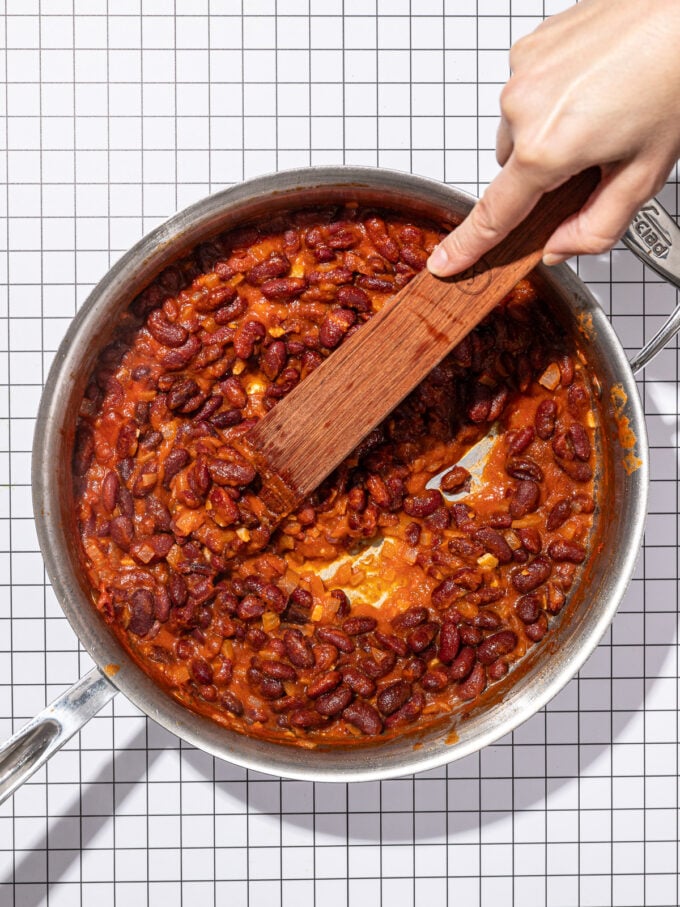 hand and wooden spoon stirring saucy kidney beans in stainless steel skillet