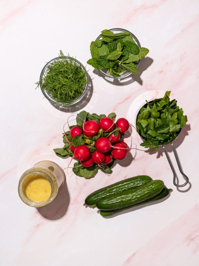 bunch of radishes, persian cucumbers, bowls of fresh herbs and lemon dressing on pink background