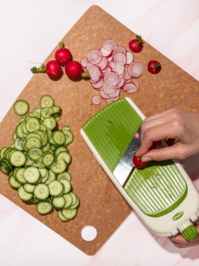 hand slicing radishes on a mandolin on chopping board with cucumbers