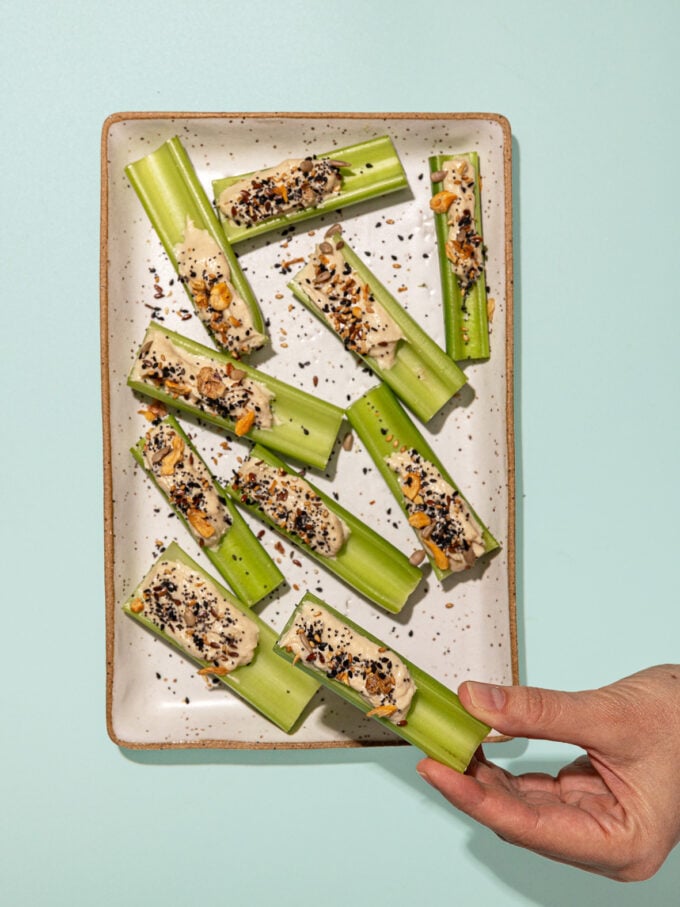 hand holding tahini stuffed celery stick next to rectangular plate of stuffed celery with seeds on top
