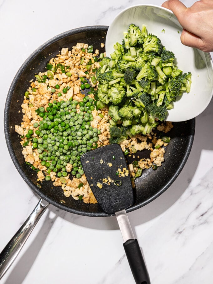 pouring bowl of chopped broccoli into skillet with frozen peas and crumbled tofu