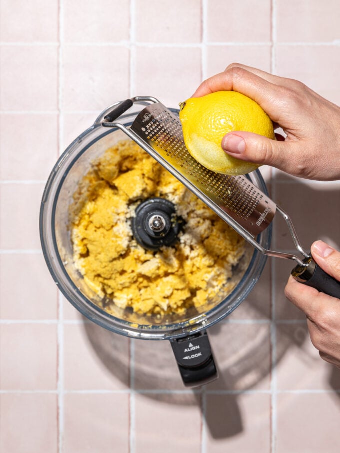 hand grating lemon zest over food processor with tofu and nutritional yeast