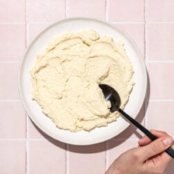 hand scooping tofu ricotta on white plate with black spoon on pink tile background