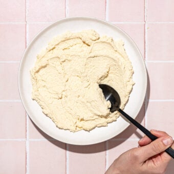 hand scooping tofu ricotta on white plate with black spoon on pink tile background