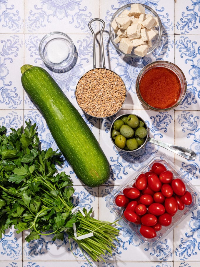 parsley, zucchini, cherry tomatoes and green olives on a white and blue tile background with a measuring cup of dry farro and bowl of vegan feta