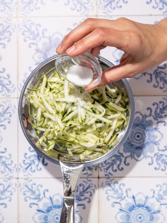 hand pouring salt onto shredded zucchini in a colander
