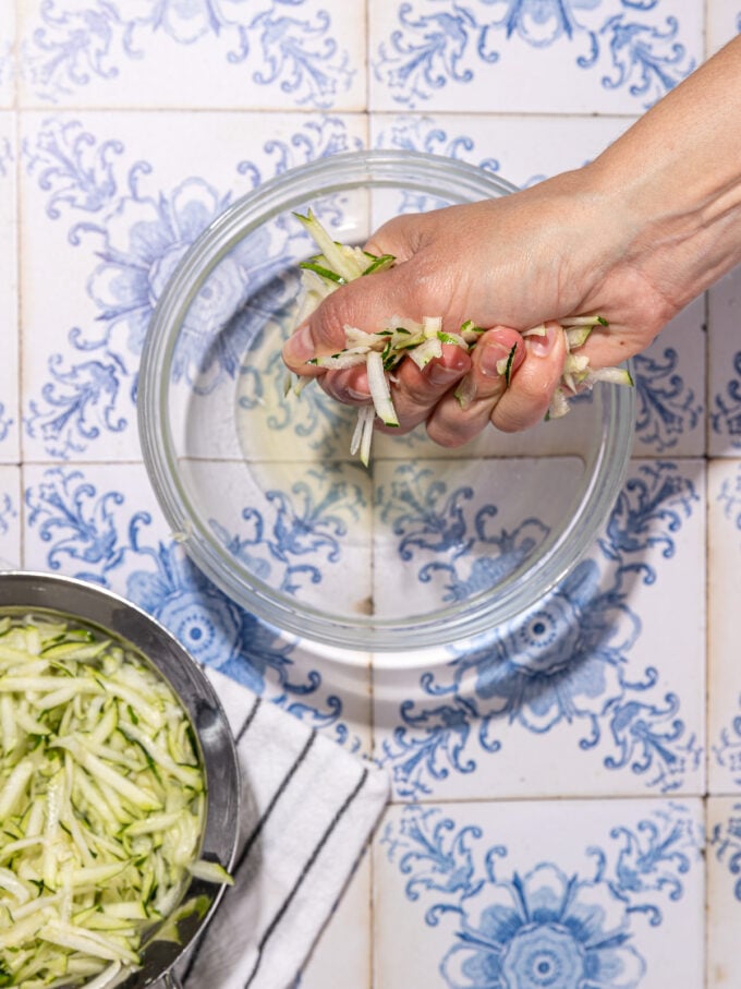 hand squeezing excess water out of a handful of shredded zucchini, over a glass bowl