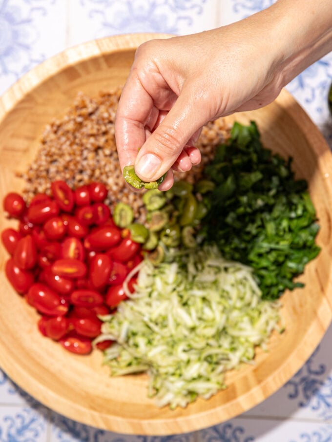 hand smashing green olives between fingers, over a wooden bowl filled with zucchini, parsley, tomatoes and farro.