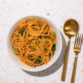 white bowl with peanut butter noodles and greens with a copper spoon and fork beside it