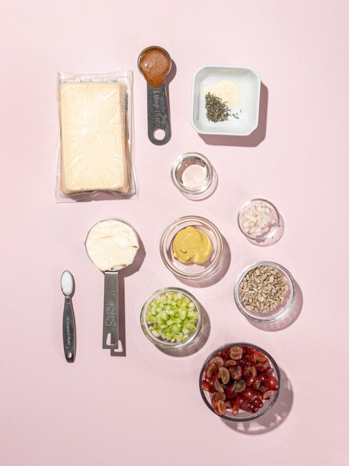 Divers bols contenant des cuillères à soupe d'ingrédients - raisins rouges, tofu, mayonnaise, céleri, bouillon - sur fond rose