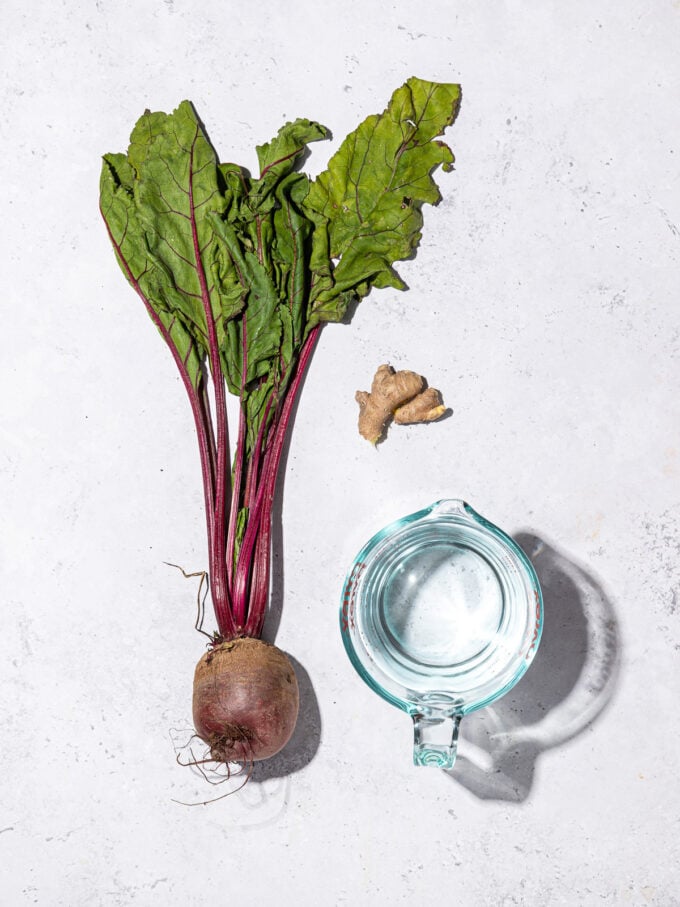 a beet with greens beside a piece of ginger and a cup of water