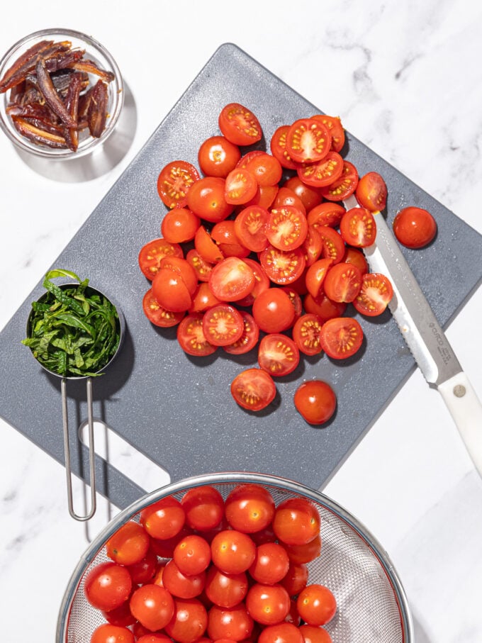 sliced cherry tomatoes on black cutting board with cup of sliced mint and cup of sliced dates beside it