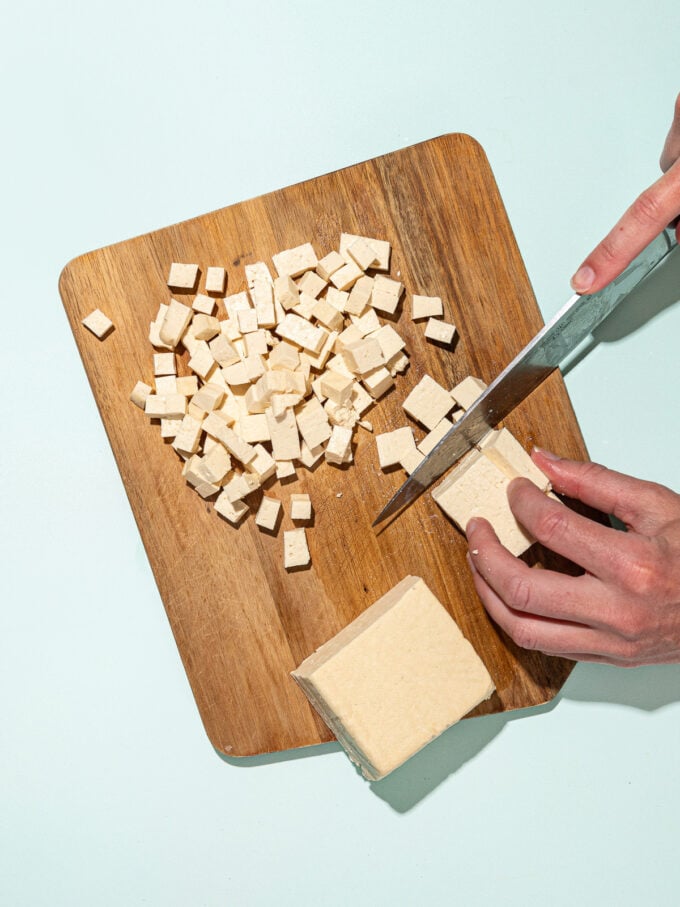 hand dicing tofu with knife on wooden cutting board