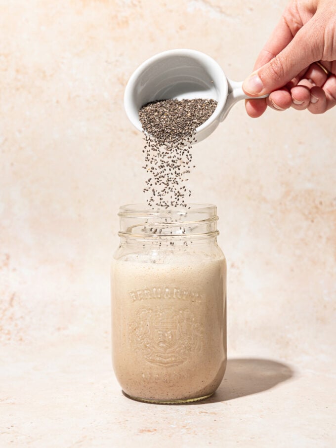hand pouring cup of chia seeds into a mason jar filled with vanilla cream