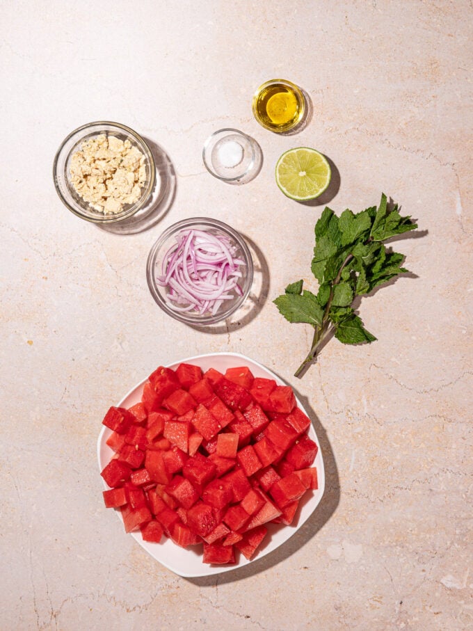 white bowl of cubed watermelon beside small bowls of vegan feta, sliced red onion, oil, salt and a lime