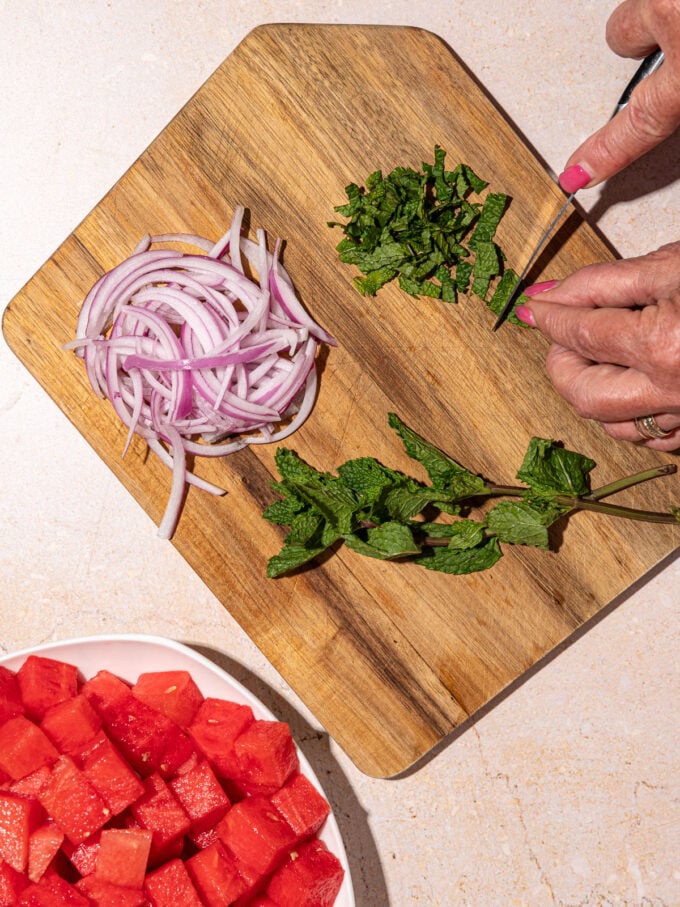 hand slicing mint leaves on wooden cutting board with sliced red onion beside it