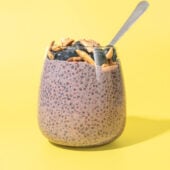 glass of blueberry lemon chia pudding with spoon tucked in, against a yellow background.