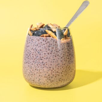 glass of blueberry lemon chia pudding with spoon tucked in, against a yellow background.