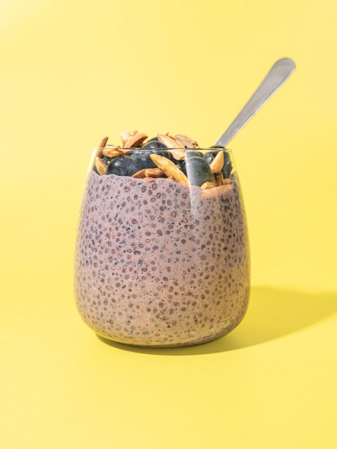 glass of blueberry lemon chia pudding with spoon tucked in, against a yellow background.