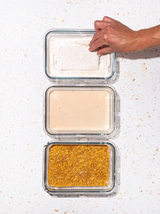 hand dipping tofu into dish of cornstarch with dishes of milk wash and cornflakes beside it