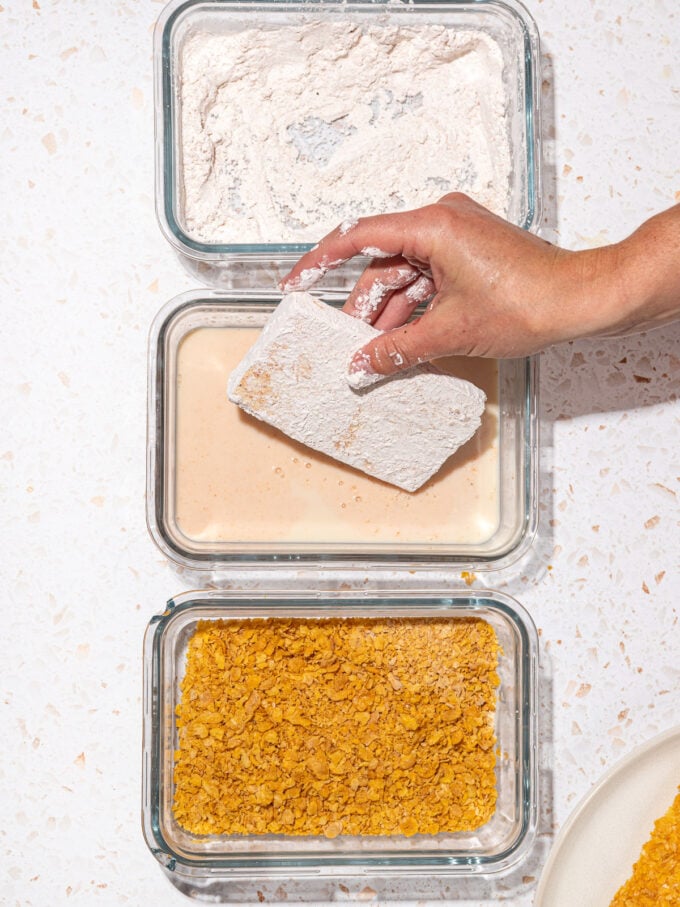 hand dipping tofu into milk wash with containers of cornstarch and cornflakes beside it in a row