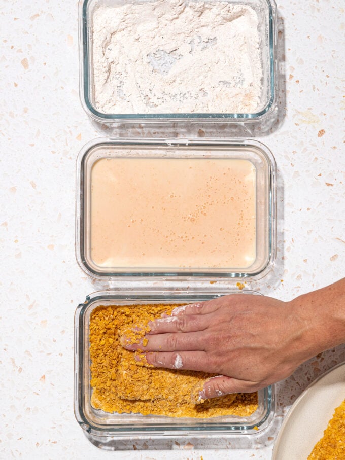 hand patting tofu into cornflakes, with dishes of cornstarch and milk wash in a row beside it