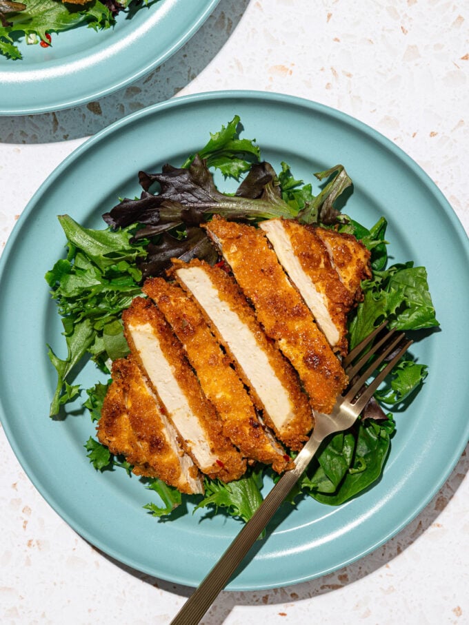 sliced tofu cutlet on bed of salad greens on top of blue plate with black fork