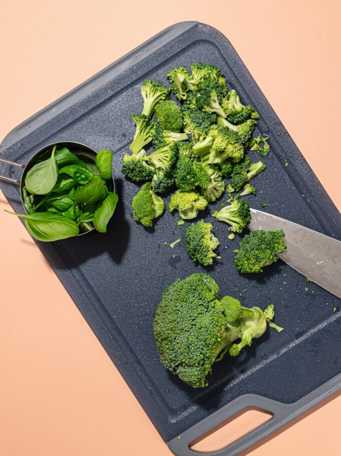 chopped broccoli with knife on black cutting board with a measuring cup full of basil leaves beside it