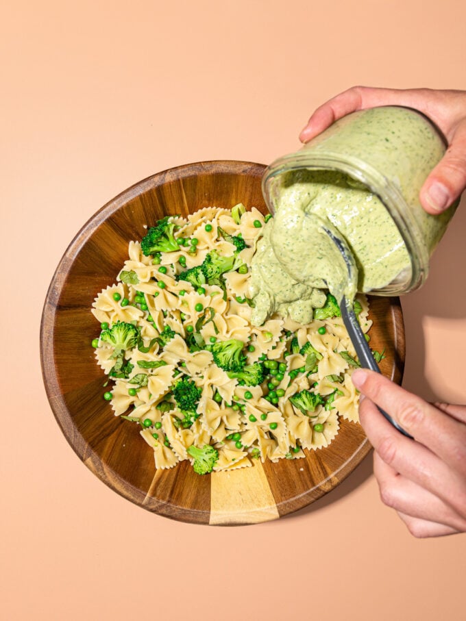 hand pouring jar of green goddess dressing into wooden bowl of pasta and broccoli