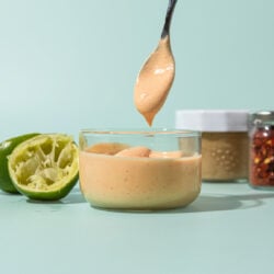 hand holding spoon of cashew dressing over glass jar of dressing, with spent limes, and jars of cashew butter and chile flakes beside it