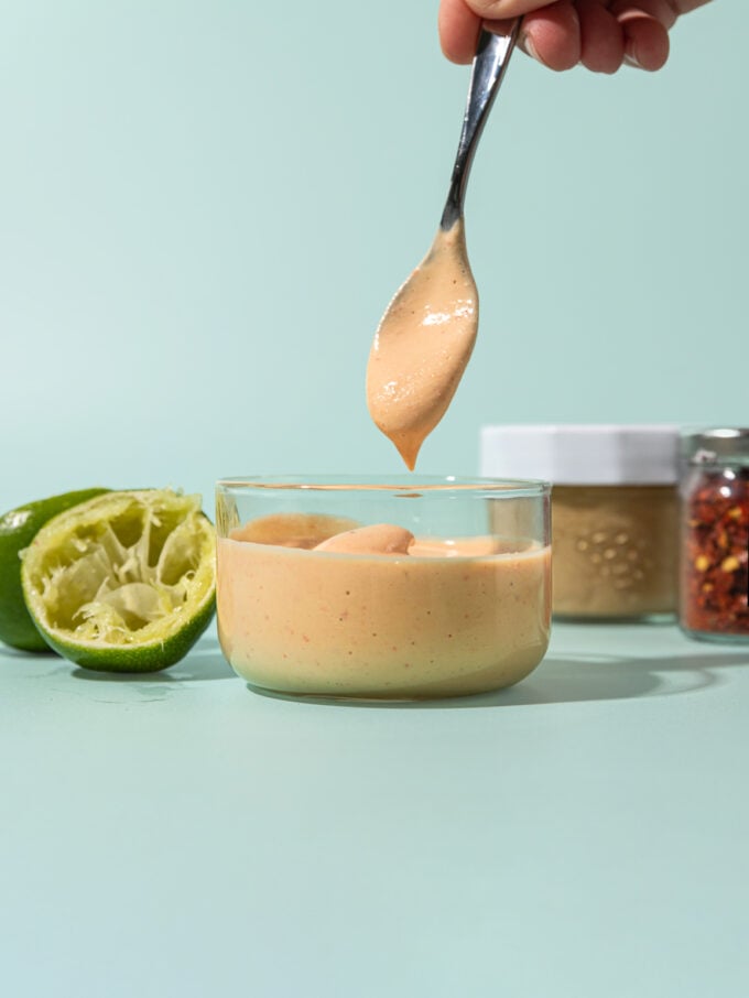 hand holding spoon of cashew dressing over glass jar of dressing, with spent limes, and jars of cashew butter and chile flakes beside it