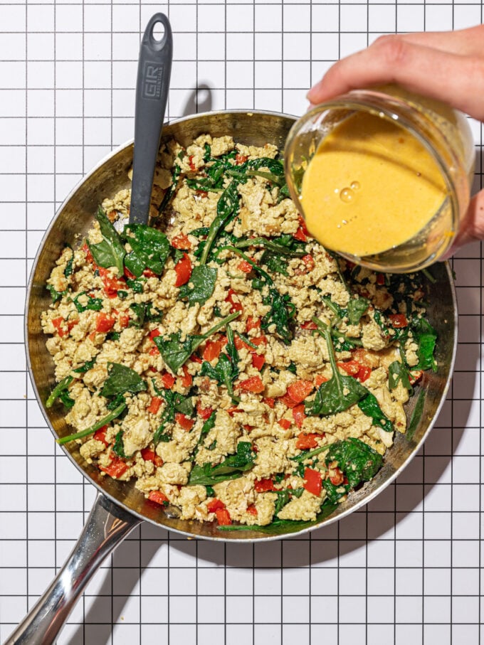 hand pouring sauce into skillet with crumbled tofu, spinach and red pepper