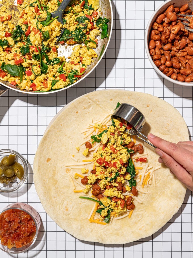 hand adding tofu scramble to tortilla with pinto beans and vegan cheese shreds