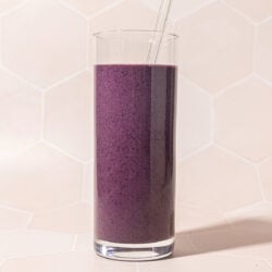 tall glass of blueberry beet smoothie with glass straw on a pink tile background