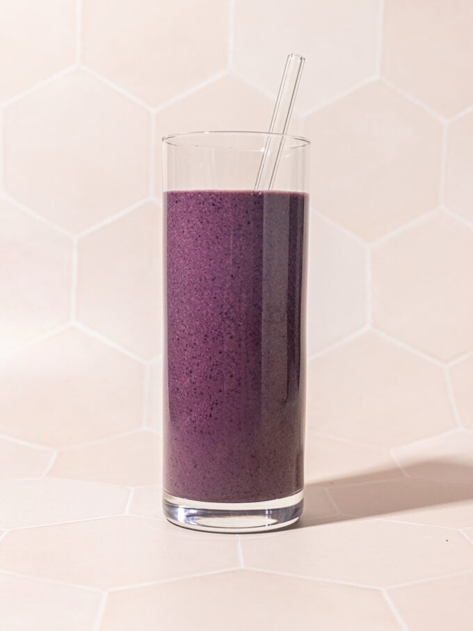 tall glass of blueberry beet smoothie with glass straw on a pink tile background