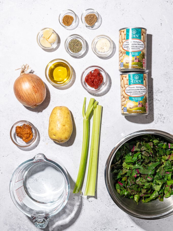 cans of white beans with bowls of ingredients: greens, tomato paste, cumin, thyme, potato, onion, celery