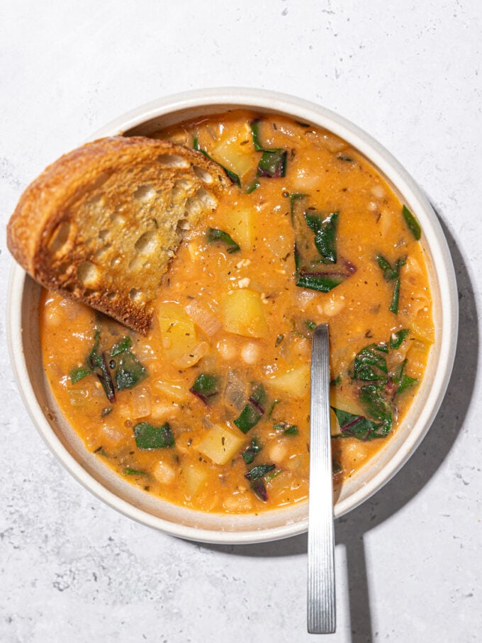 white bean stew in a white bowl with a spoon and slice of toast dunked in