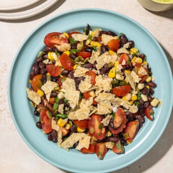 black bean corn tomato salad on blue bowl with stack of white plates to the side, and a small jar of avocado dressing