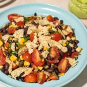 black bean, corn and tomato salad in blue bowl, with glass jar of avocado dressing behind it and a small stack of white plates
