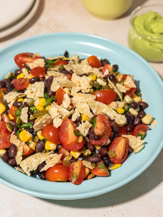 black bean, corn and tomato salad in blue bowl, with glass jar of avocado dressing behind it and a small stack of white plates